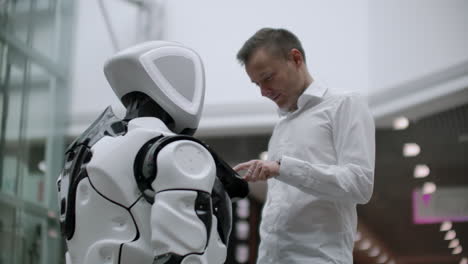 a man in a shirt communicates with a white robot asking questions and pressing the screen with his fingers