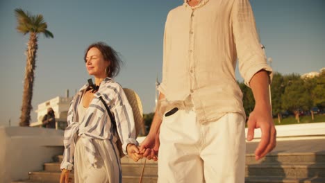 Close-up-shot-of-happy-couple-guy-and-girl-walking-on-modern-beach-holding-hands-in-beachwear-in-summer.-A-guy-in-light-clothes-and-a-brown-haired-girl-go-down-the-stairs-to-the-beach