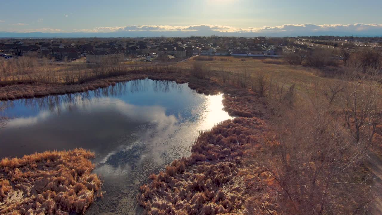 Backing up in the late afternoon sun over trees to reveal cloud reflections on a pond below with the Rocky Mountains yonder.  Shot in Greeley Colorado