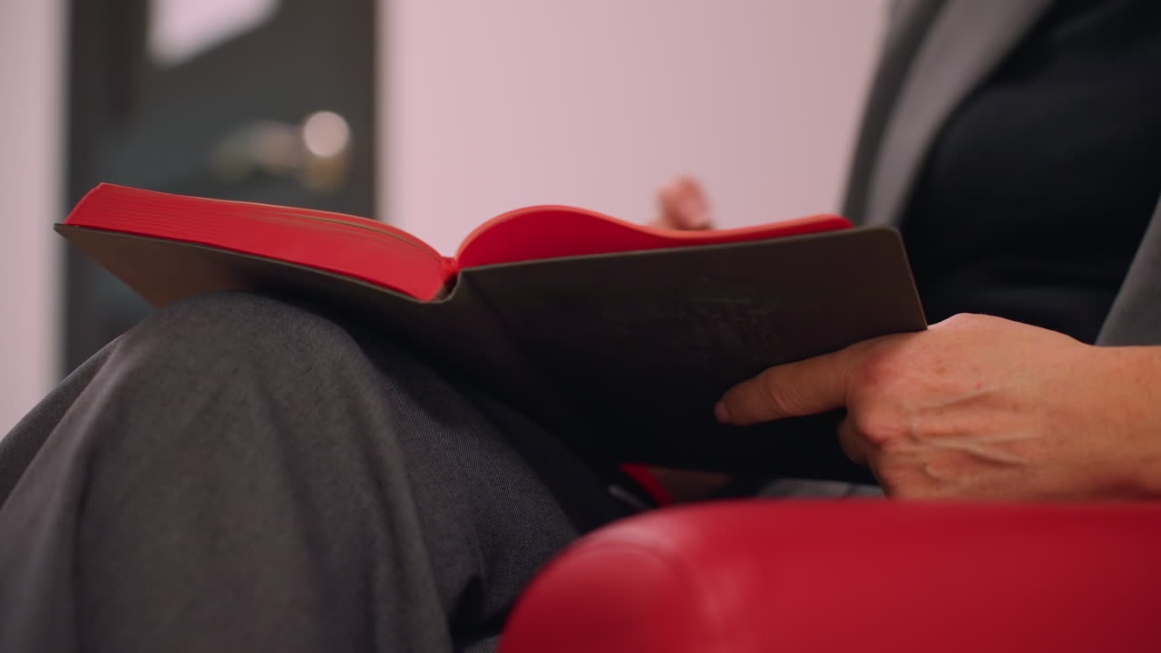 Close-up of businesswoman's hands holding notebook with red cover, sitting on red armchair, reading and reviewing notes, preparing for important meeting or planning session