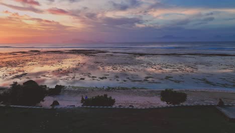 Descending-drone-clip-of-beautiful-multicolored-sunset-reflecting-on-calm-sea-and-coral-reef,-in-Alas-Purwo-National-Park,-Java,-Indonesia