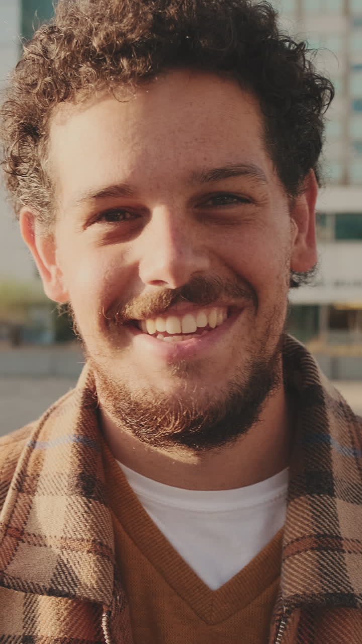 A man with curly hair and a plaid shirt smiles outdoors