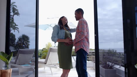 expecting couple standing together, smiling and holding pregnant belly on balcony