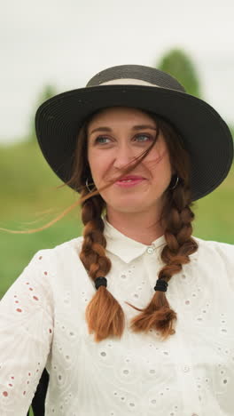 una mujer con un vestido blanco y un sombrero negro está sentada en un campo verde, sonriendo cálidamente. el entorno al aire libre destaca su expresión alegre, un momento de felicidad en un entorno natural
