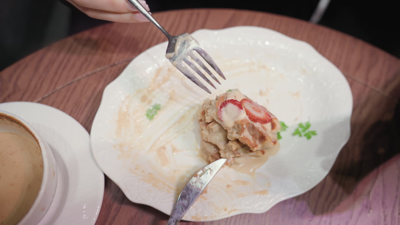 Close view of remaining creamy strawberry waffles on white plate with used knife beside latte cup on wooden table. Scene captures casual dining moment after enjoying delicious dessert in cozy setting
