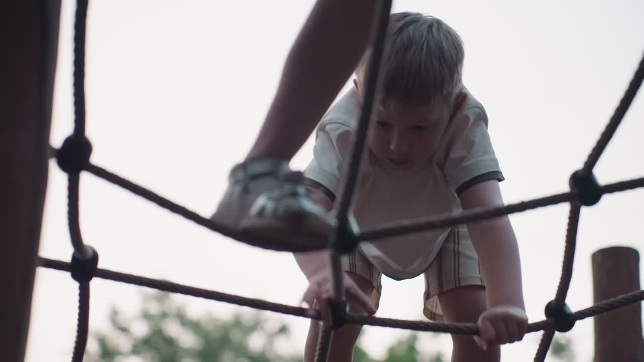 playful children carefully climbing rope net structure in park playground at dusk, capturing concentration and teamwork as they navigate climb test under soft evening light among trees