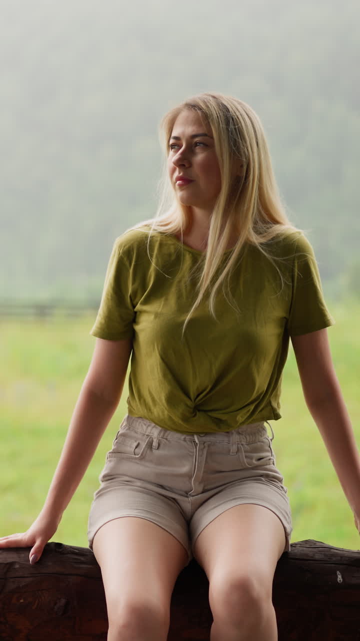 Thoughtful lady in green t-shirt enjoys nature beauty even at drizzling rain sitting on log fence near lush meadow and mountain silhouette slow motion