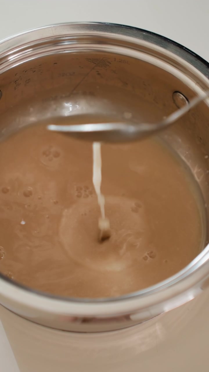 Primo piano dall'alto di salsa cremosa marrone all'interno di una pentola in acciaio inossidabile, mentre un cucchiaio di metallo mescola e un sottile getto di liquido si versa al centro, formando schizzi, increspature concentriche, bolle e una consistenza lucida
