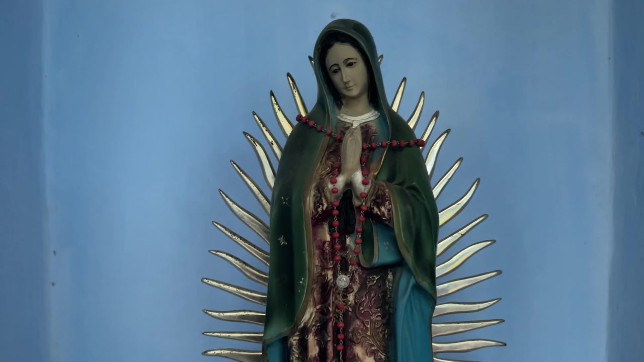 Primo piano di una statua religiosa della Vergine Maria decorata con rosari, contro una parete blu, all'interno di un ambiente spirituale.