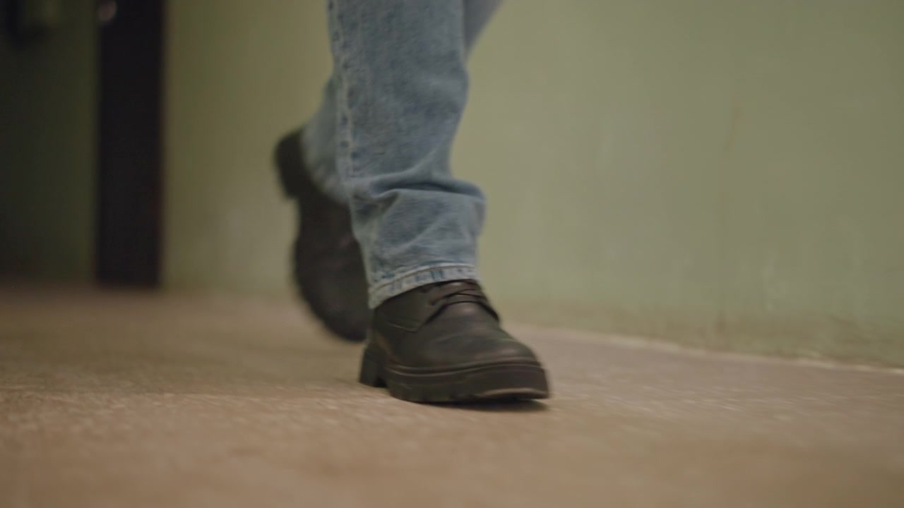 Low angle indoor view of person walking across hard floor toward doorway, showing blue denim jeans and worn black shoes in mid stride, with right foot forward, left foot lifting and subtle heel lift