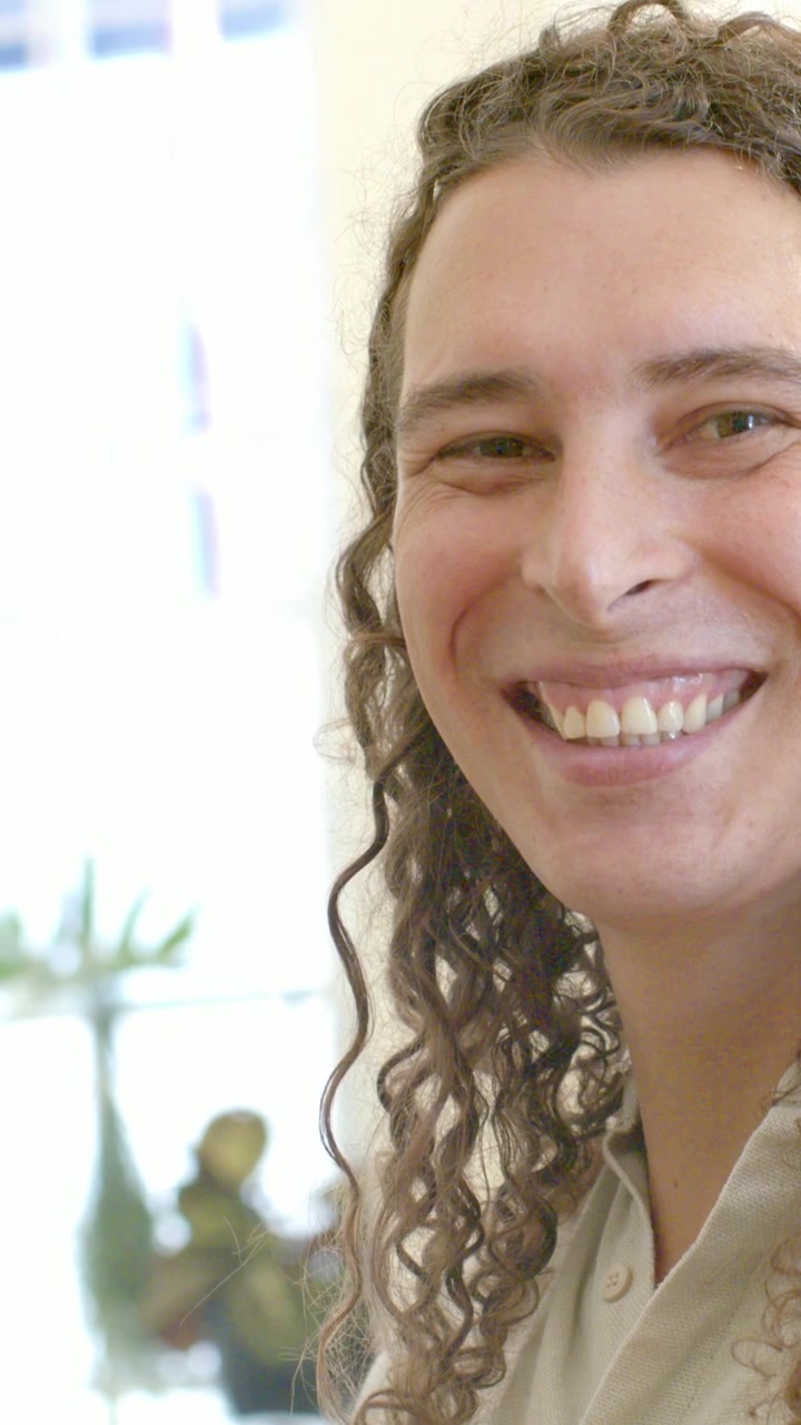 Video vertical: Cámara alejándose mostrando a una persona con camisa beige cuidando plantas en una ventana, sonriendo.