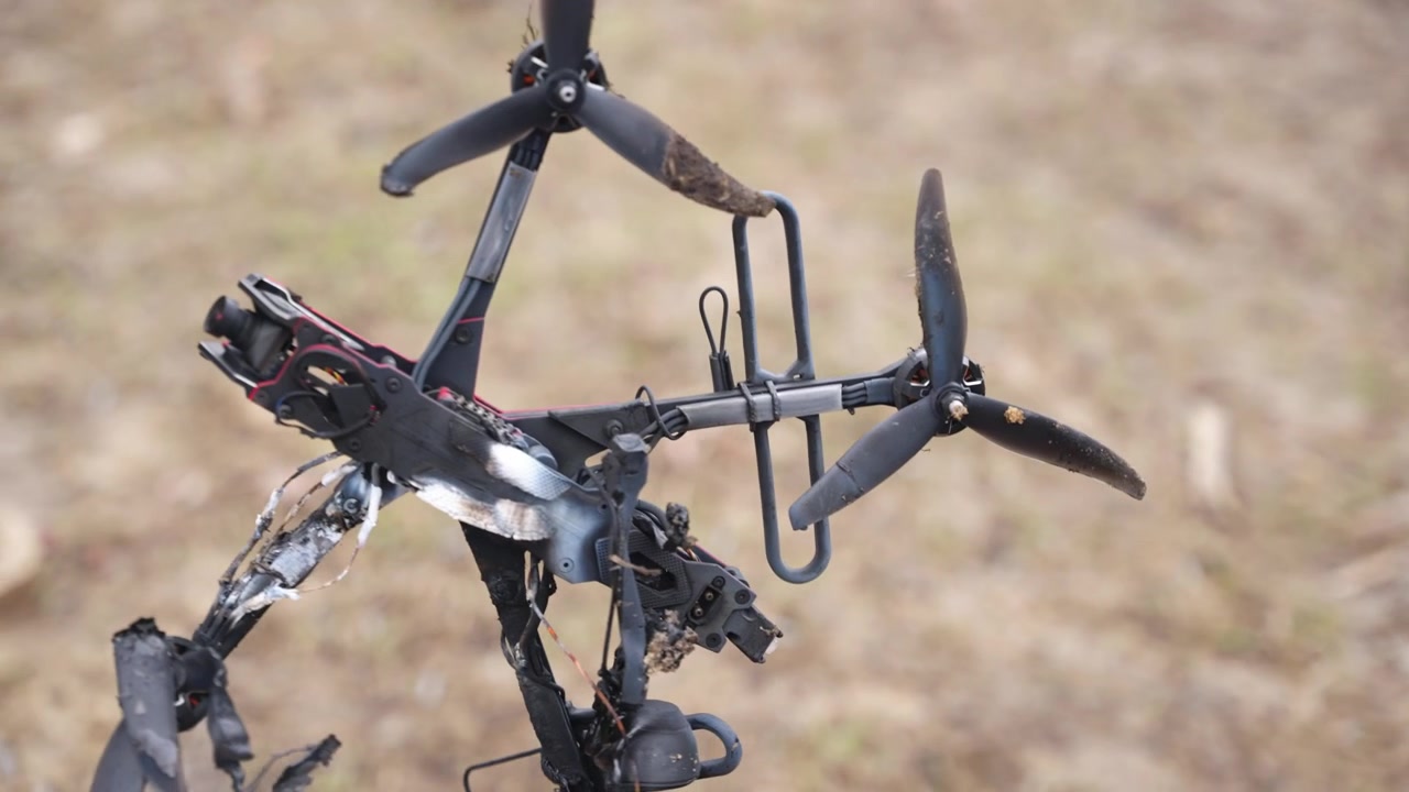 Orang memegang rangka drone FPV yang terbakar dengan kerusakan yang terlihat dan komponen yang terbuka di luar ruangan.