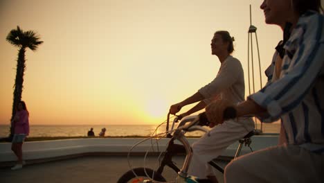 side view of a happy couple, a guy in light clothes and a girl with a straw hat ride bicycles along the beach near the sea at sunrise in summer. a guy and a girl are driving along the beach at dawn