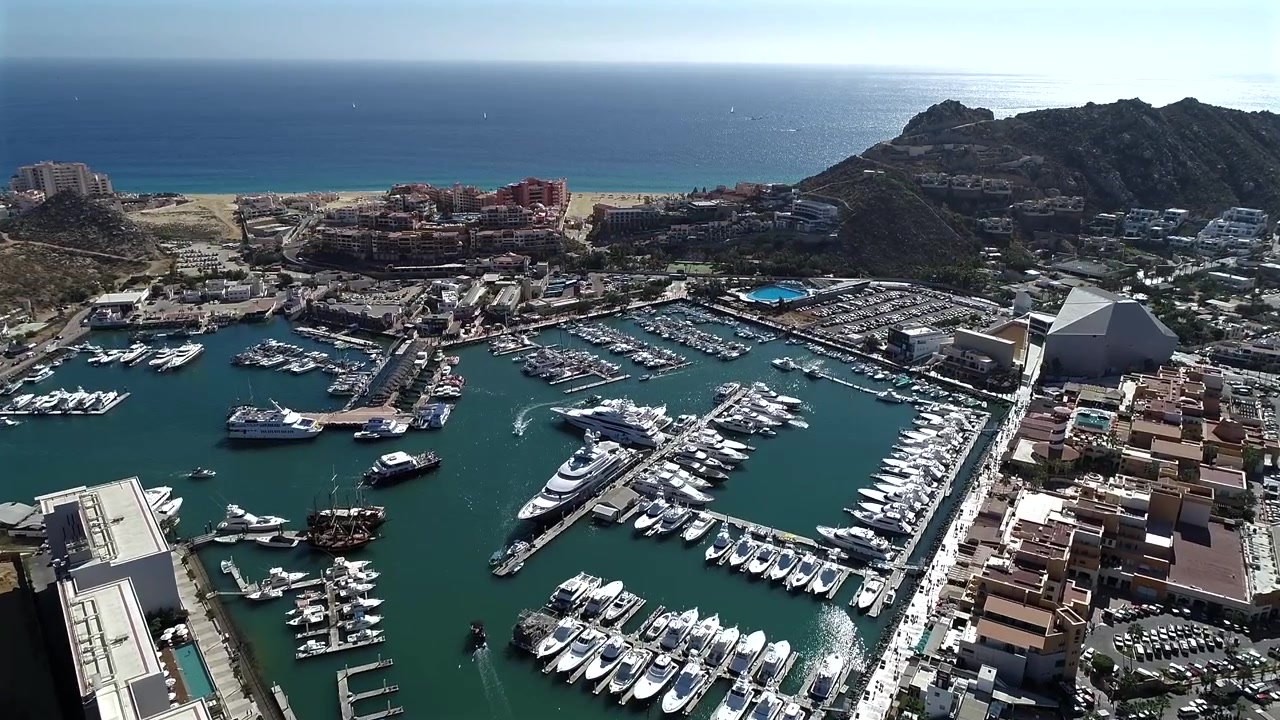 Drone olha para baixo em um ângulo de aproximadamente 45 graus sobre a marina de Cabo San Lucas, movendo-se por um porto cheio de iates e barcos, passando por hotéis e sobre um resort de luxo à beira-mar