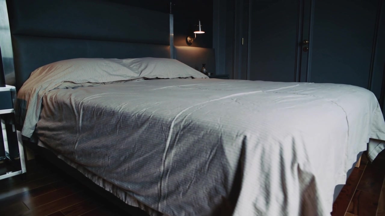 Mouvement panoramique de la caméra commençant vers la droite à travers la chambre, révélant le lit, l'oreiller, la lampe et la porte fermée.