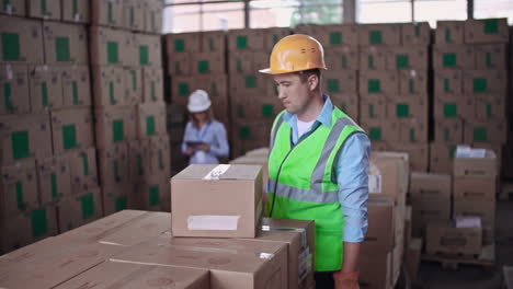 Trabajador-De-Almacén-Con-Casco-Y-Chaleco-Reflectante-Sella-Una-Caja-Con-Cinta-Adhesiva,-En-El-Fondo-Borroso-Una-Mujer-Está-Colocando-Cajas-De-Cartón-En-El-Estante