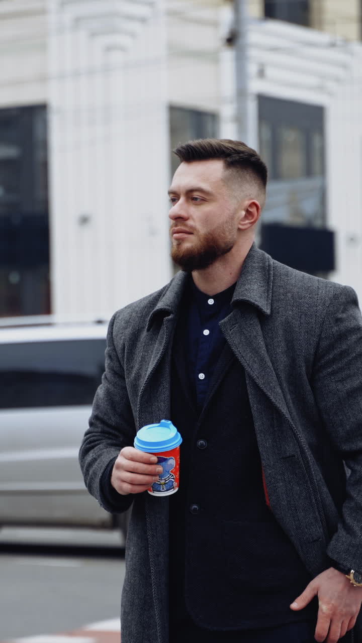 Handsome man in the city. Young businessman waiting for someone on the urban traffic background. Man in coat holds a plastic glass with coffee and looks on watch. Vertical video