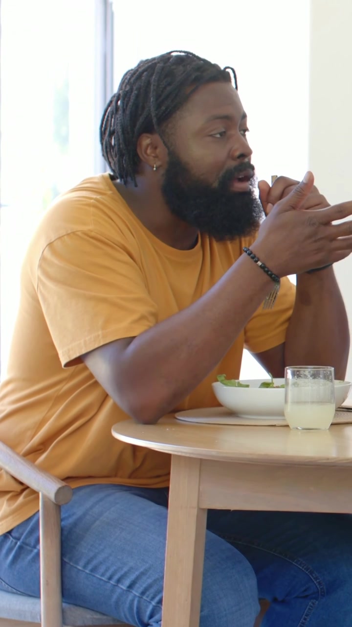 Vídeo vertical: Homem de camiseta laranja reagindo a um estímulo fora do enquadramento, comendo salada com garfo em casa.