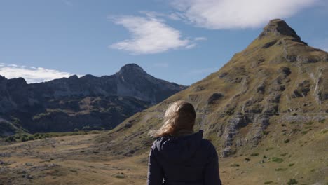 woman with softly wind-blown hair stands facing a pointed mountain in durmitor national park under blue sky
