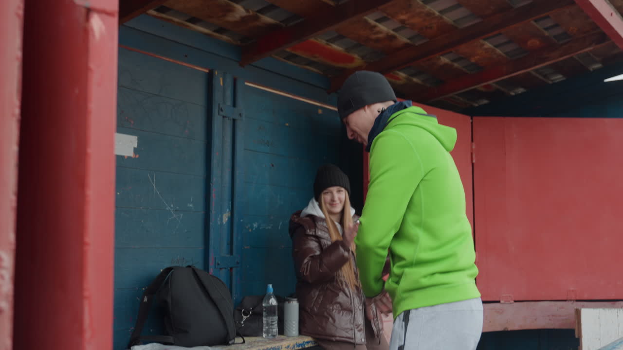 Un hombre caucásico con sudadera verde se acerca a un amigo sentado para ofrecerle agua en una estructura exterior; un gesto amistoso con una mochila y botellas en un banco; una interacción natural que muestra afecto y espíritu de equipo.
