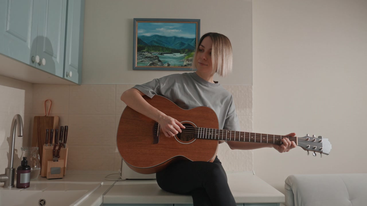 Donna bianca che suona la chitarra in cucina, pratica acustica delicata sul bancone dei mobili blu, a piedi nudi, cantautrice calma che fa le prove, atmosfera domestica intima con un quadro e un divano sullo sfondo, musica