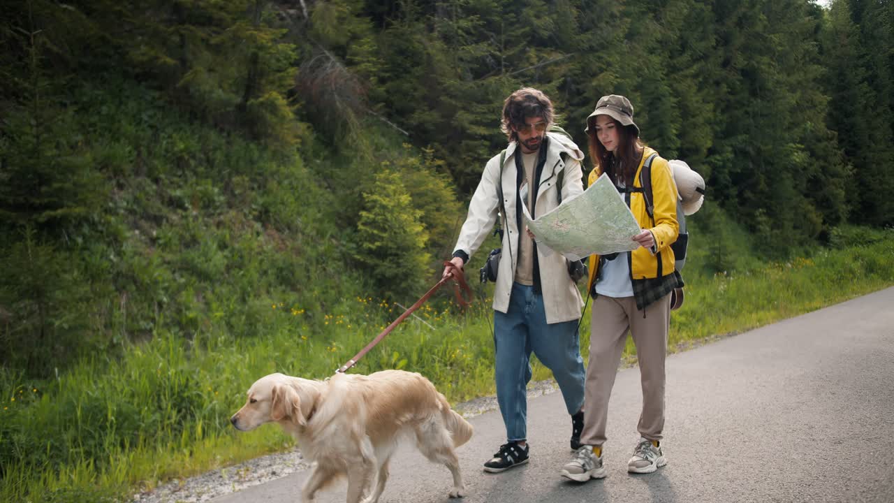 un chico y una chica turistas en ropa especial de senderismo, junto con su perro de color claro, están mirando un mapa con el fin de entender dónde van a lo largo de la carretera a lo largo del bosque