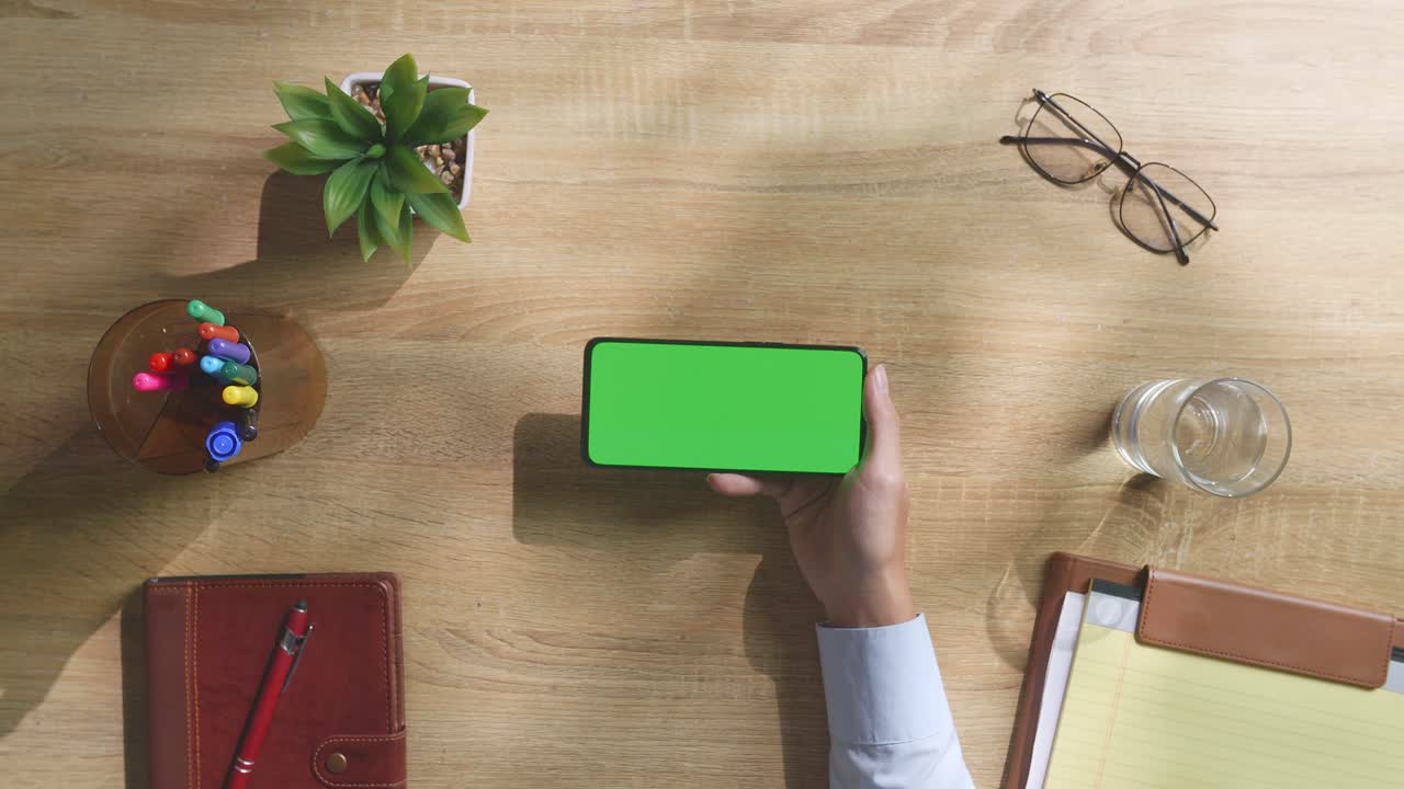 vista de arriba hacia abajo de una computadora portátil con pantalla verde simulada pantalla chromakey en un escritorio de oficina de madera al lado de la libreta con bolígrafos, gafas y un vaso de agua.