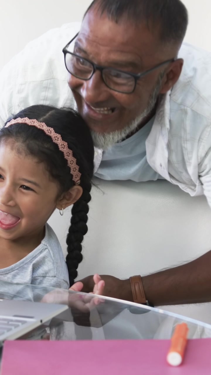 Bambina che ride dopo che un uomo maturo si china, indica e sussurra, guidando il laptop su un tavolo di casa