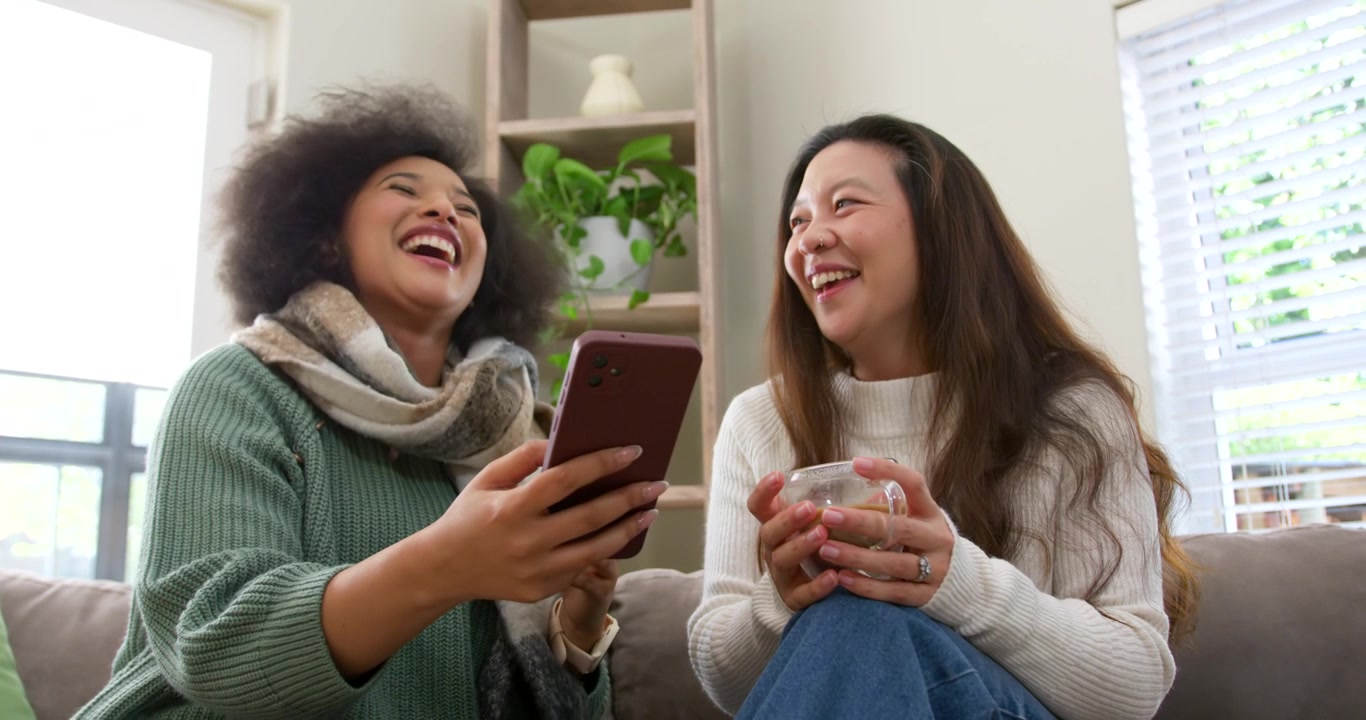 Diverse female friends in knits on couch left woman showing phone both holding glass mugs laughing