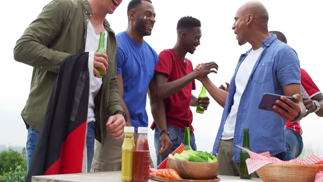 Cinque amici uomini chinati su un tavolo da picnic, un uomo che tiene uno smartphone, gli altri che brindano con la birra