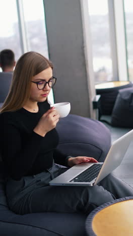 beautiful lady in office near the window. attractive young business woman working on a laptop and drinking coffee. pretty girl in comfortable place having break during work. vertical video