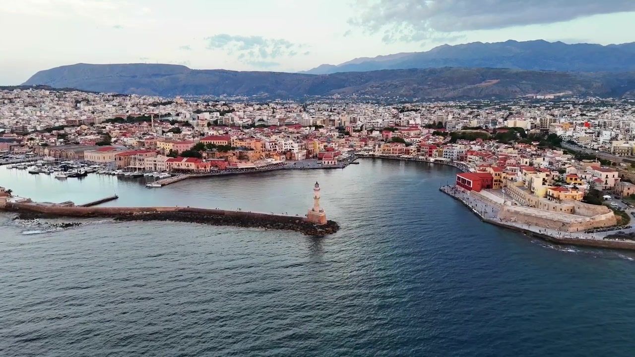 Il vecchio porto veneziano di Chania presenta il Faro Egiziano, un frangiflutti in pietra e un lungomare storico sotto le Montagne Bianche a Creta, sorvolo con drone