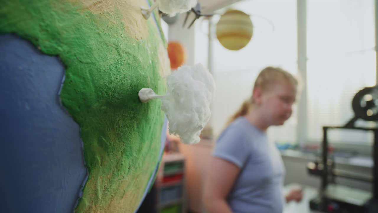 Elementary School Girl Attaching Miniature Models of Wind Turbine and Clouds to Globe