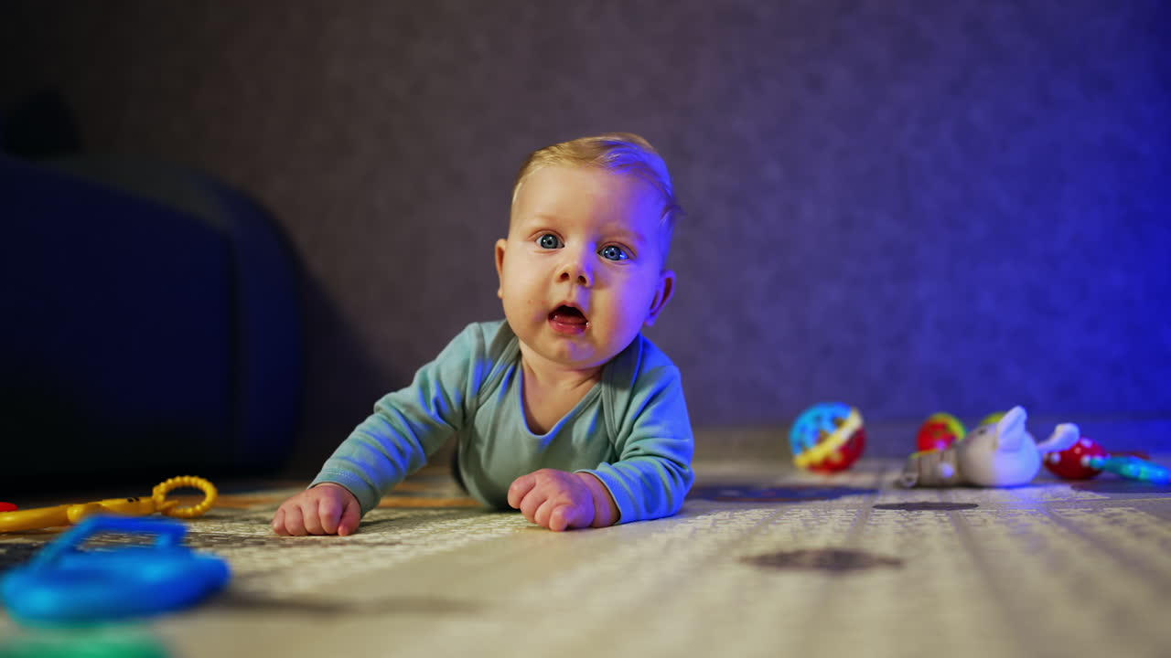 Adorable blonde baby boy lies on belly looking up with curiosity. Cute child lies at home on the floor among the toys.
