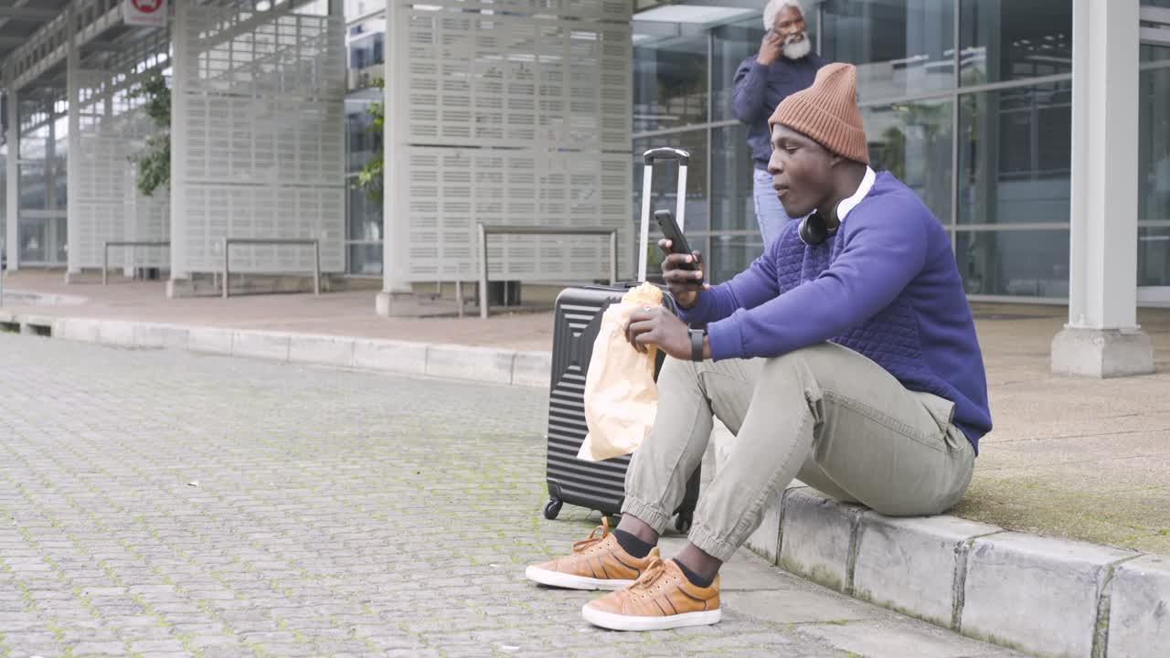 Uomo afroamericano che apre un sacchetto di snack e scorre il telefono al terminal in attesa di un passaggio.