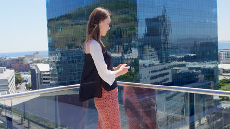 Side-view-of-young-caucasian-businesswoman-texting-on-mobile-phone-and-standing-in-office-balcony-4k