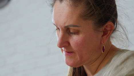 close view of woman with natural makeup wearing beige outfit and red earrings, looking downward with calm focused expression, standing against soft white brick wall background under daylight