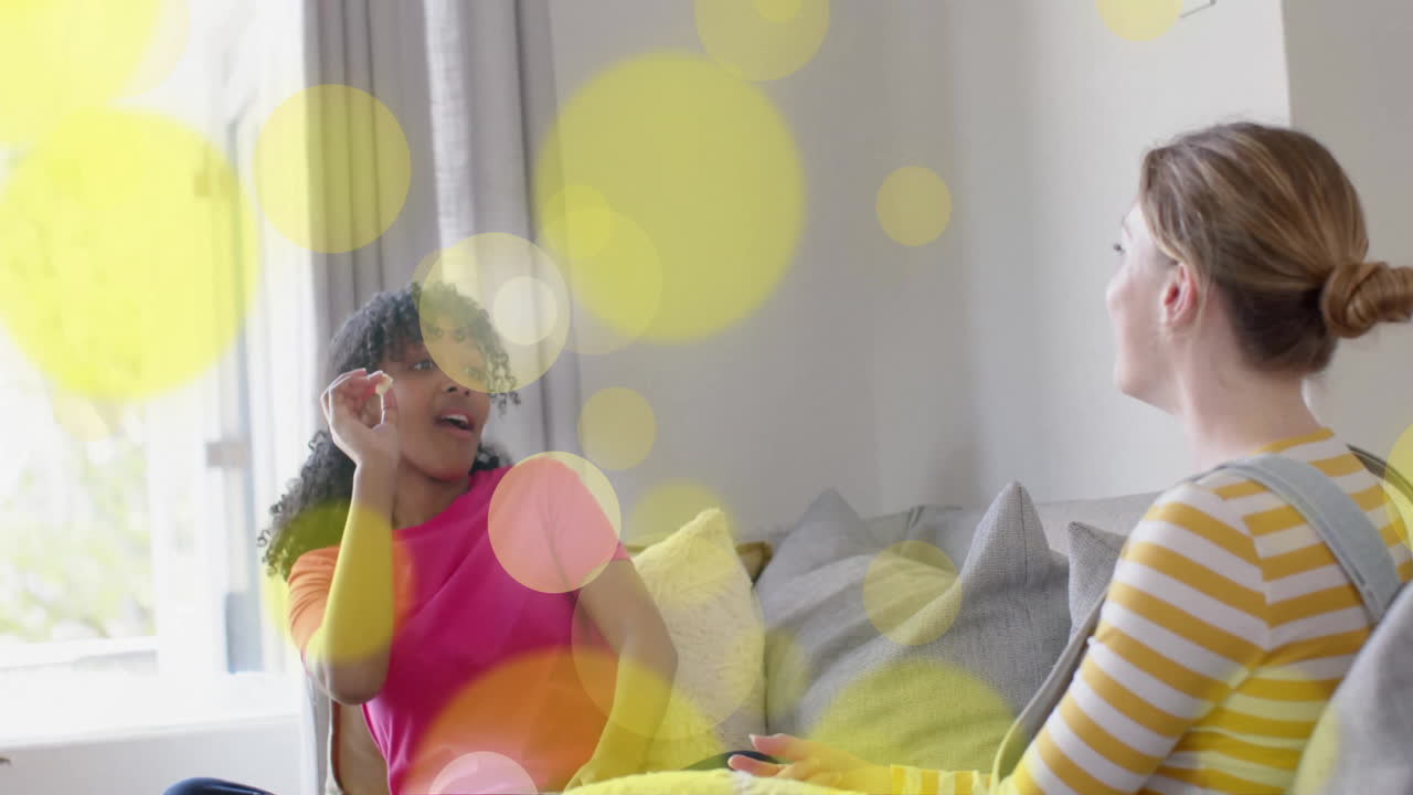 Animation of yellow light spots over two diverse teenage girls laughing and playing with popcorn