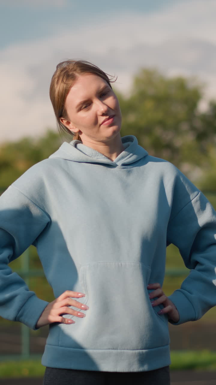 허리에 손을 고 왼쪽에서 오른쪽으로 머리를 돌리는 시안 후디를 입은 여성, 배경에는 농구 허프와 함께 다른 사람의 그림자가 그녀에 여 있습니다.