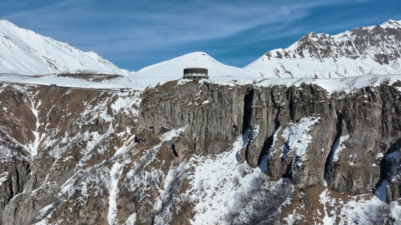 drone shot of A unique circular structure stands on the edge of a towering cliff, surrounded by snow-capped mountains. A perfect blend of human-made architecture and natural beauty