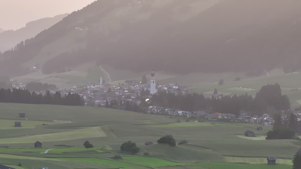 Idyllic village nestled in rolling green fields at sunrise with misty mountains in the background