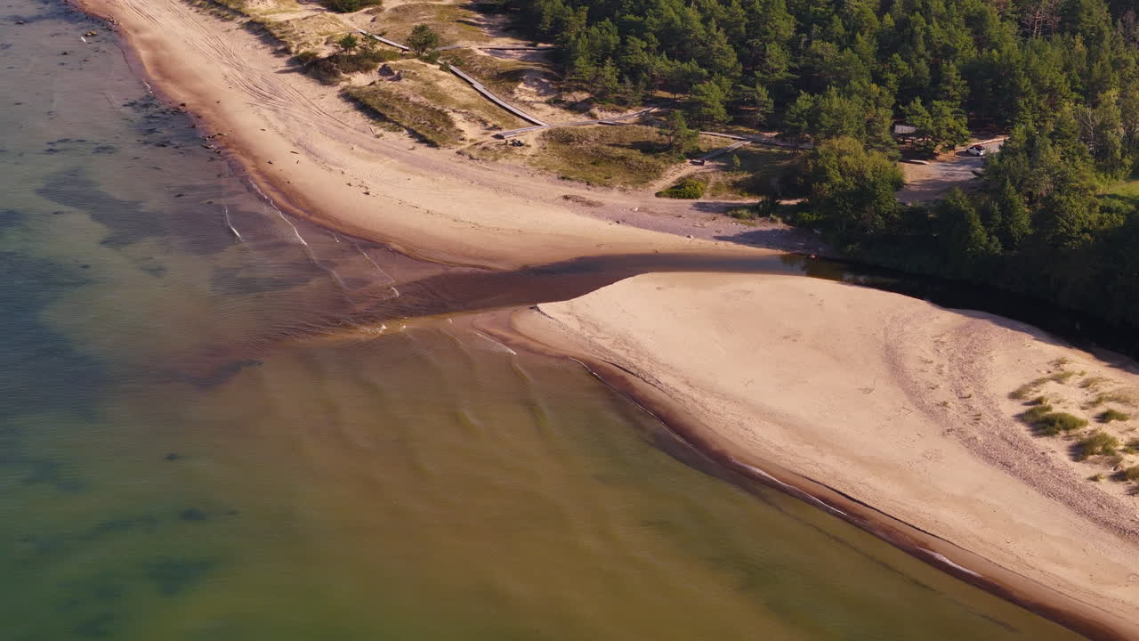 Drone follows Uzava river mouth meeting Baltic Sea with sand and greenery views