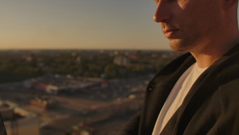 A male freelance programmer sits on a skyscraper roof with a laptop and beer typing code on a keyboard during sunset. Remote work