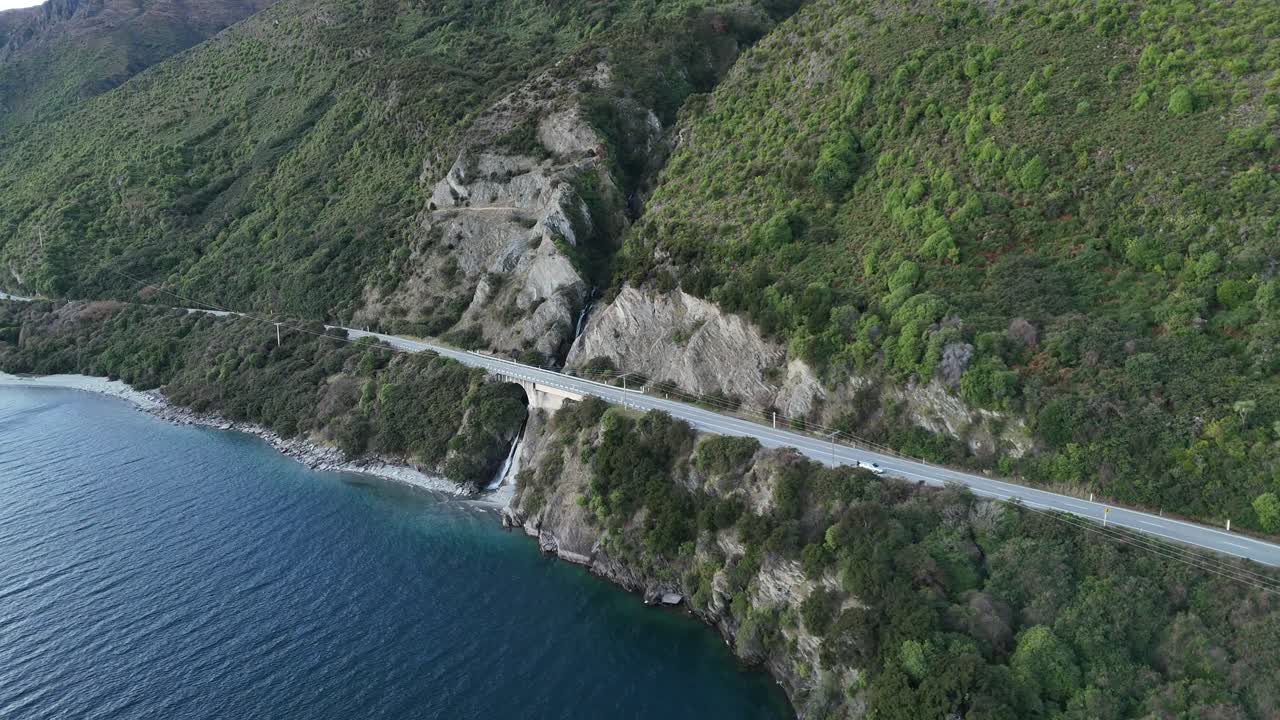 Aerial view of natural lake Hawea in New Zealand with waterfall and road coming from mountain. Approaching shot. Turquoise shoreline