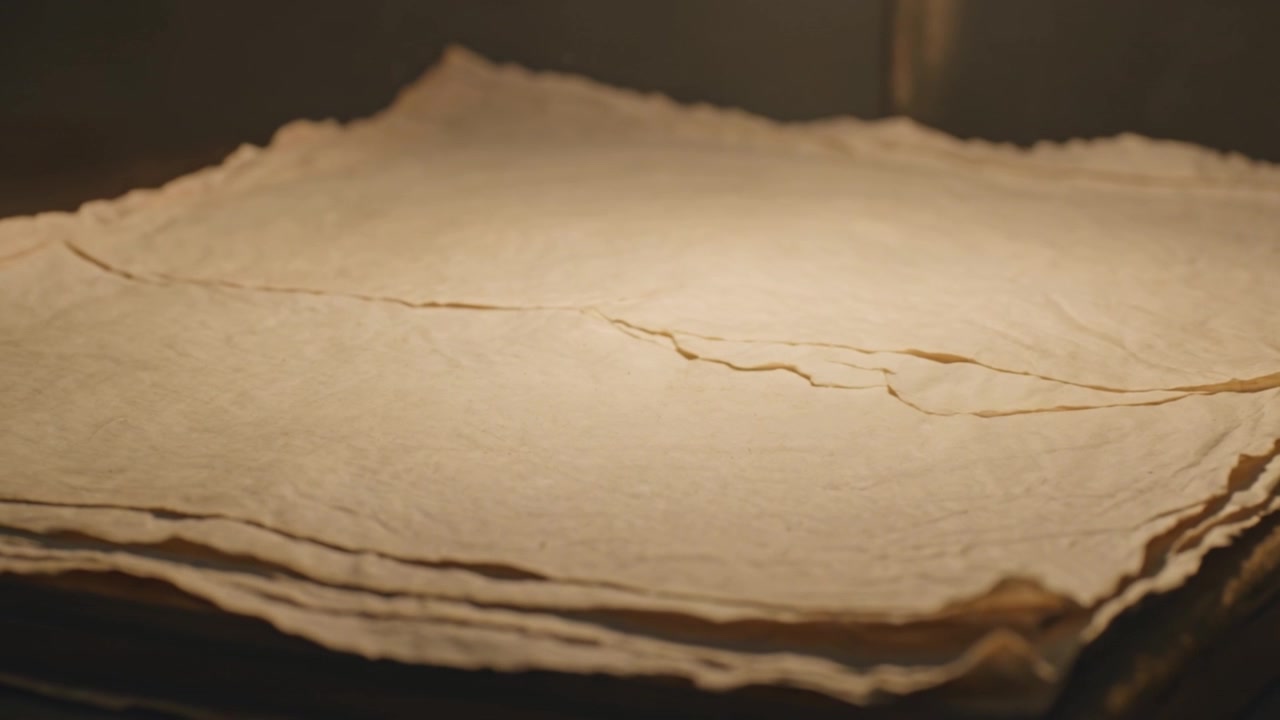 Pila di carta fatta a mano illuminata su una scrivania scura, con luce calda e messa a fuoco variabile che rivela la grana e le pieghe.