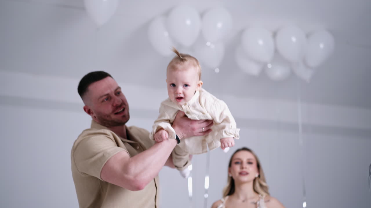 Caucasian man waving his little baby boy in hands approaching him to camera. Smiling mother stands at backdrop. Studio footage.