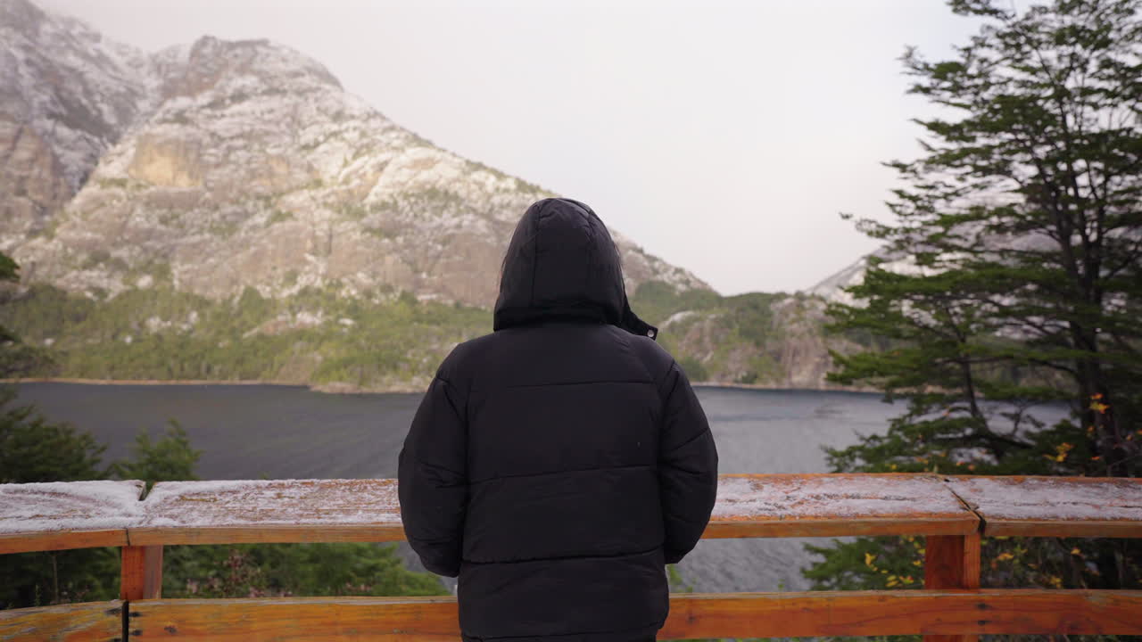 Slow panning clip frames solitary figure against the vast mountain peaked panorama and lake’s surface down on scene. Ideal for introspection or travel brand narratives