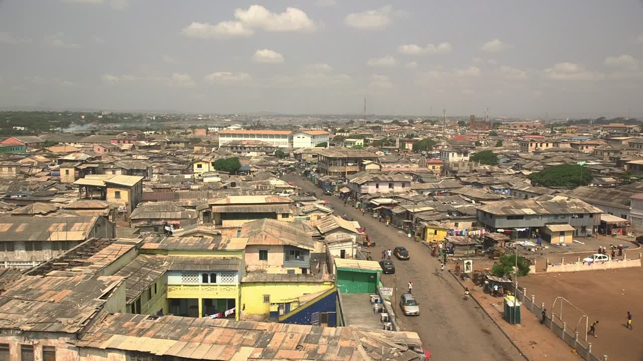 Aerial view of a densely populated neighborhood in Accra, Ghana, showing corrugated rooftops, busy streets, and everyday urban life. shops lining the streets