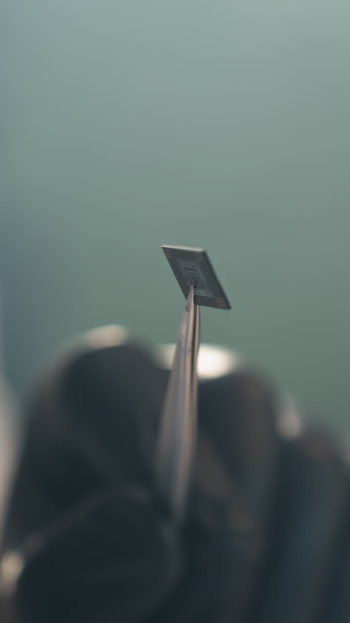 A close-up view of a technician's gloved hand holding a small electronic component with tweezers under a microscope