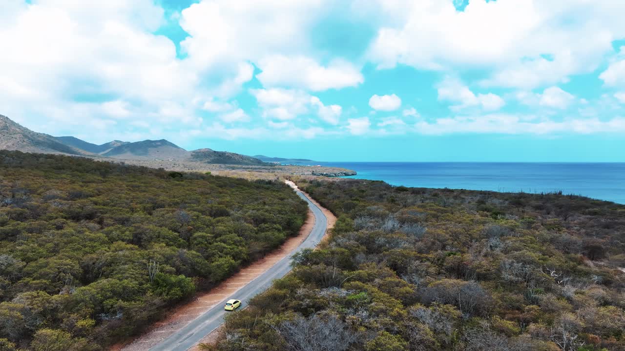 드론은 쿠라카오의 바다 근처의 작은 도로에서 자연을 통해 운전하는 노란색 자동차를 따라가고 있습니다.